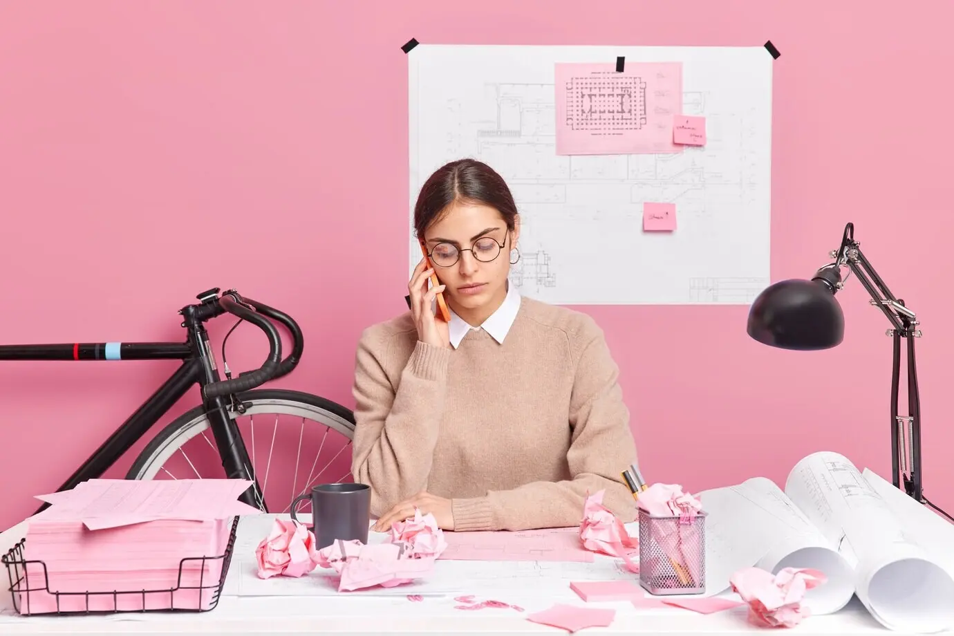 Junge talentierte Architektin fertigt Skizzen zur Gebäudeplanung an, führt ein Telefongespräch, ist in den Arbeitsprozess eingebunden und posiert am Schreibtisch, umgeben von Papieren und Bauplänen.