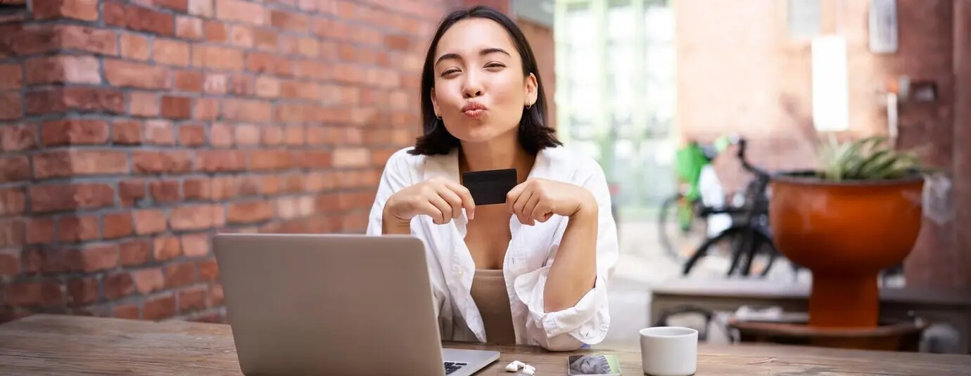 Porträt eines jungen, stylischen Mädchens, das neben einem Laptop sitzt, eine Kreditkarte zeigt und lächelt, Rechnungen bezahlt und online einkauft.